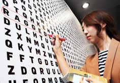 a woman is writing on a wall with letters and numbers behind her she is holding a clipboard