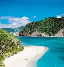 an island with white sand and blue water surrounded by green hills on the side of it