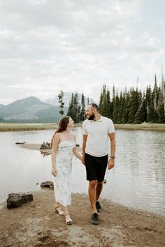 a man and woman standing next to each other near a body of water with trees in the background