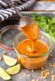 a spoon full of peanut sauce being lifted from a glass bowl with limes on the side