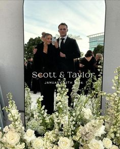 a man and woman standing in front of a mirror with flowers on the ground next to it