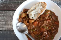 a white bowl filled with stew and bread on top of a wooden table next to a spoon