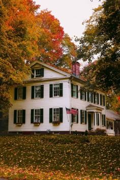 a large white house with green shutters on the front and side windows surrounded by fall foliage