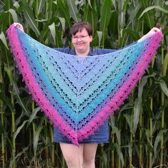 a woman standing in front of a corn field holding up a blue and pink crocheted shawl