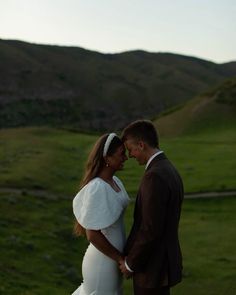 a man and woman standing next to each other in front of a green field with hills