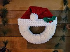 a knitted santa claus hat hanging on a wooden wall with pine cones and evergreens