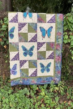 a blue butterfly quilt hanging on a fence in front of some green plants and trees