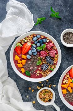 two bowls filled with fruit and granola on top of a gray surface next to spoons