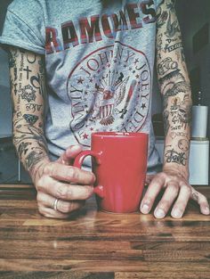 a man with tattoos holding a red coffee mug on top of a wooden table in front of him