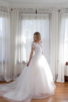 a woman in a white wedding dress is standing by a window with sheer drapes