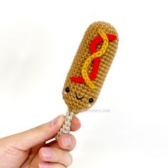 a hand holding a crocheted hot dog on a stick with mustard and ketchup