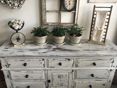 an old white dresser with potted plants on top and two clocks hanging from the wall