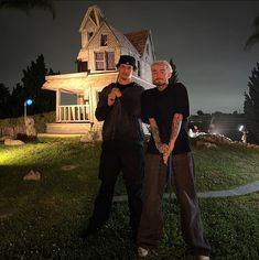 two men standing next to each other in front of a house at night with the lights on