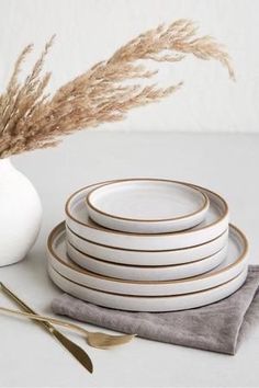 a stack of white plates sitting on top of a table next to a vase filled with dry grass