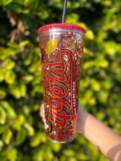 a person holding up a cup with a lid and straw in front of some bushes