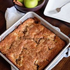 a square cake in a pan on a table with apples and cinnamon sticks next to it