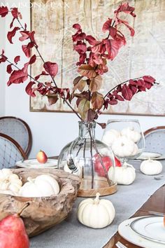 the table is set with white pumpkins and red leaves in a glass vase on it