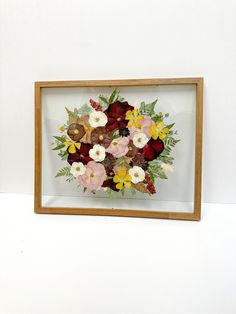 an arrangement of flowers in a wooden frame on a white wall behind it is a vase with red, yellow and pink flowers