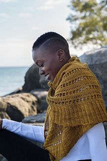 a woman sitting on rocks next to the ocean using her cell phone while wearing a yellow knitted shawl