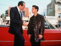 two men shaking hands in front of a red car