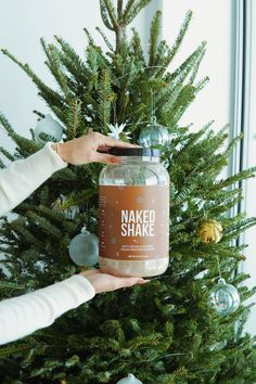 a woman holding up a jar of naked shake next to a christmas tree