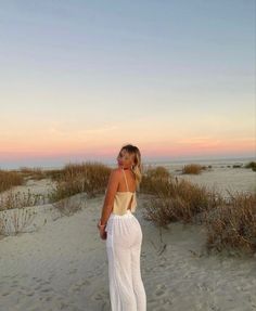 a woman standing on top of a sandy beach next to tall grass and bushes at sunset