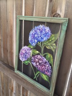 a painting of purple and blue flowers in a green frame hanging on a wooden fence