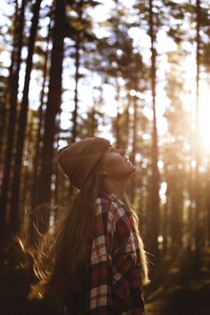 a woman with long hair standing in the woods looking up at the sun shining through the trees