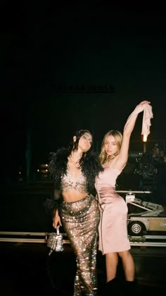 two women standing next to each other in front of a car with their arms up