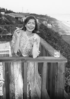 two women sitting on a deck near the ocean and holding drinks in their hands, one woman is smiling at the camera