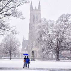 two people are standing under an umbrella in the snow near a large building and trees