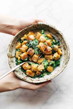 two hands holding a bowl of food with broccoli and chicken in it on a marble surface