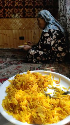 a white plate topped with yellow rice next to a woman sitting on a couch in the background