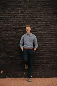 a man leaning against a brick wall with his hands in his pockets and smiling at the camera