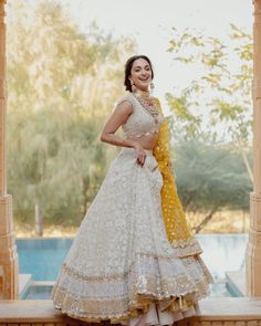 a woman in a white and yellow lehenga standing on a ledge near a pool