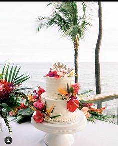 a wedding cake with tropical flowers and greenery sits on a table near the ocean