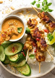 chicken skewers with rice, cucumber and sauce on a plate next to sliced cucumbers