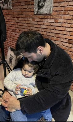 a man holding a small child in his lap while sitting on a chair next to a brick wall