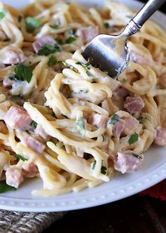 a white plate topped with pasta covered in ham and parmesan cheese next to a fork