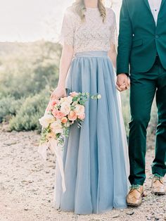 a man and woman holding hands walking down a dirt road with flowers in their hand