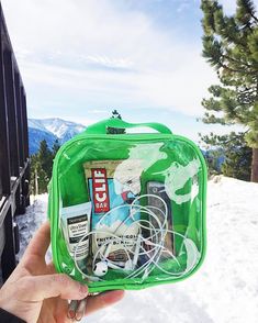 a person holding up a green bag with various items inside it in the snow near some trees