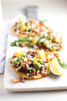 three tacos on a white plate with lime wedges