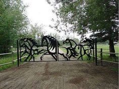 a metal gate with horses on it in the middle of a dirt road next to trees