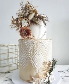 a white and gold wedding cake decorated with dried flowers, leaves and feathers on a marble table