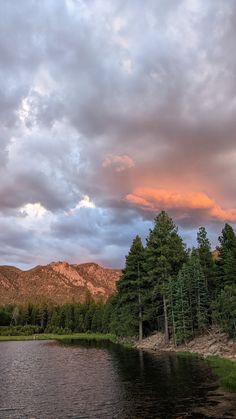 a lake surrounded by trees and mountains under a cloudy sky with pink clouds in the distance