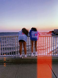 two girls are looking out over the water from a pier at sunset or sunrise with their backs turned to the camera