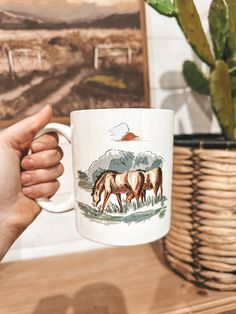 a person holding a coffee mug with horses on it in front of a potted plant