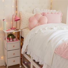 a white bed with pink pillows on top of it next to a night stand and nightstand