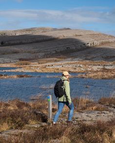 a woman with a backpack is walking by the water and hills in the distance,