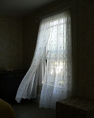 a bed sitting next to a window covered in white sheer curtains and a night stand